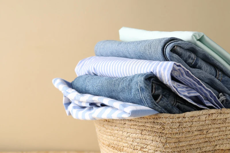 Neatly folded shirts and jeans stacked in a woven wicker laundry basket