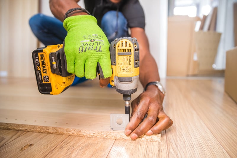 Gloved hands using a yellow power drill to drive a screw into a metal bracket on wood flooring